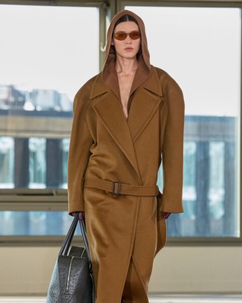 Zdjęcie z kompanii Stella McCartney z kolekcji wiosna-jesień 2025. Przedstawia ekologiczne trendy w modzie premium: modelka jest ubrana w komplet ze sztucznego zamszu. Ma na sobie brązowy płaszcz z kapturem i paskiem na biodrach. W ręce niesie ciemnobrązową torebkę, a na nosie ma okulary przeciwsłoneczne.
