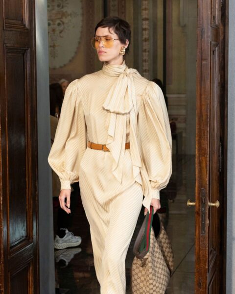 Zdjęcie z pokazu marki Gucci. Modelka ma na sobie żółtą, plisowaną sukienkę z bufiastymi rękawami i wiązaniem pod szyją. W talii pasek, na nosie okulary przeciwsłoneczne, a w ręce torebka typu shopper. Modelka ma krótkie, brązowe włosy.