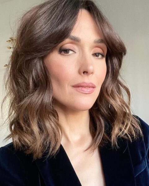 Na zdjęciu aktorka Rose Byrne w marynarce z granatowego weluru. Jej makijaż jest naturalny i podkreśla oczy w odcieniach brązu i różu. Włosy, koloru kasztanowego, są ułożone w swobodne fale, a twarz otulają delikatne pasma, tworząc stylową grzywkę curtain bangs.