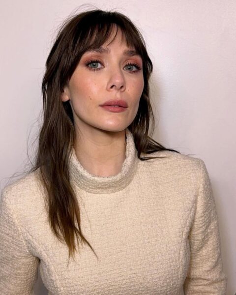 Na zdjęciu aktorka Elizabeth Olsen w swetrze z golfem w odcieniu ecru. Na jej twarzy widać delikatny, naturalny makijaż z wyraźnie zaznaczonymi oczami za pomocą cieni w odcieniach różu i brązu. Jej długie, brązowe włosy są lekko pofalowane, a na czoło opada postrzępiona, lekka grzywka.