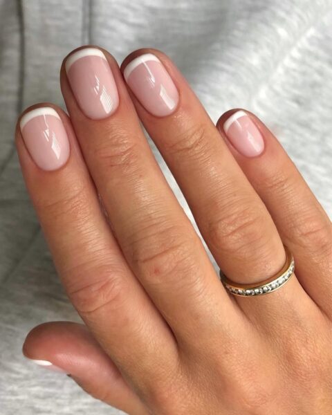 Zdjęcie dłoni ze zbliżeniem na krótkie, owalne paznokcie z delikatnym, lśniącym french manicure. Na palcu serdecznym widać pierścionek z brylancikami.