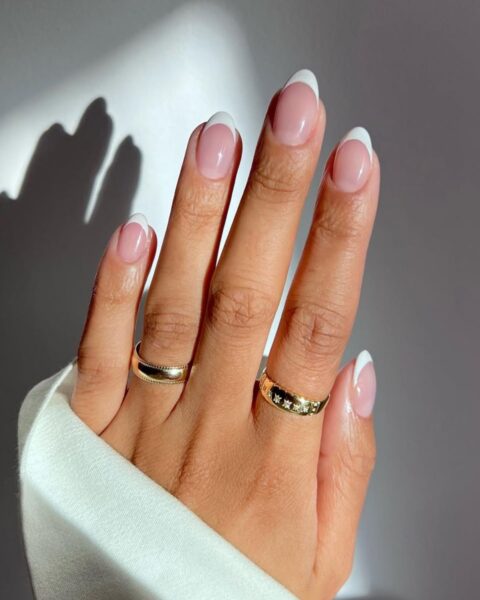Zdjęcie dłoni z długimi, owalnymi paznokciami z french manicure. Na palcach widać dwa złote pierścionki.
