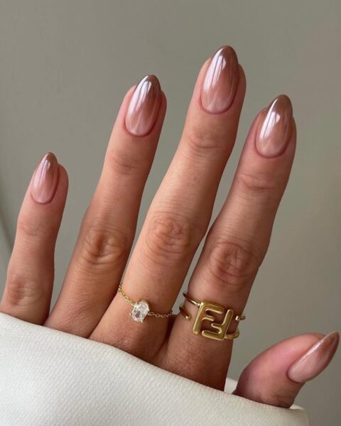 Zbliżenie na dłoń z eleganckim manicurem ombre (gradient) w odcieniach beżu i brązu. Długie, migdałowe paznokcie mają gładkie przejście koloru, zaczynając od jasnej, mlecznej bazy przy skórkach, aż do połyskujących, głębokich brązowych końcówek. Na palcach widoczne są dwa złote pierścionki: delikatny z kryształkiem oraz masywniejszy z motywem geometrycznym. Dłoń jest częściowo zakryta białym materiałem.