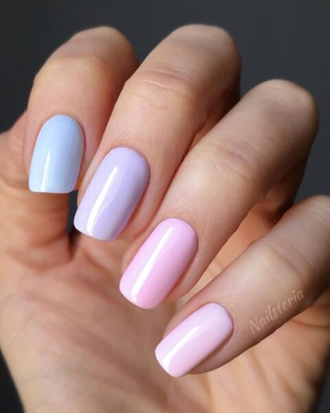 Zbliżenie na dłoń z wiosennym manicurem skittles (gradient) w pastelowych kolorach. Krótkie, kwadratowe paznokcie pomalowane są różnymi odcieniami: od jasnego błękitu na kciuku, przez lawendowy fiolet, po dwa odcienie pastelowego różu na pozostałych palcach. Manicure jest błyszczący, a tło ciemne, co podkreśla jasne kolory paznokci.