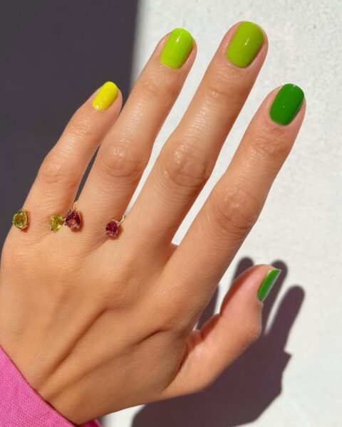 Zbliżenie na dłoń z manicurem gradientowym (skittles), w którym każdy paznokieć jest pomalowany innym odcieniem zieleni i żółci. Kolory przechodzą od jasnego, intensywnego żółtego na palcu serdecznym, przez limonkową zieleń, aż do nasyconej, trawiastej zieleni na palcu wskazującym. Dłoń nosi złoty, otwarty pierścionek ozdobiony trzema różnokolorowymi kamieniami szlachetnymi w odcieniach zieleni, żółci i różu.