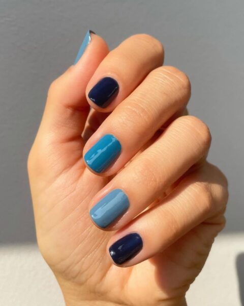 Zbliżenie na dłoń z manicurem gradientowym (skittles) w odcieniach niebieskiego. Krótkie paznokcie, opiłowane na kształt kwadratowy, są pomalowane różnymi kolorami: od bardzo ciemnego granatu na małym palcu i palcu wskazującym, przez stonowany błękit/szaroniebieski, aż do jasnego, nasyconego turkusu na palcu środkowym. Dłoń jest oświetlona jasnym światłem słonecznym.