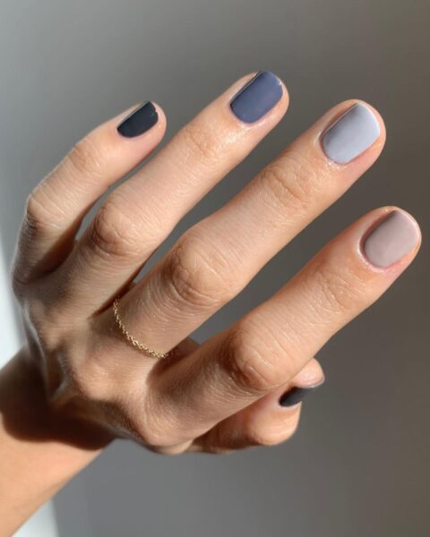 Zbliżenie na dłoń z manicurem gradientowym (skittles) w stonowanych, chłodnych odcieniach. Krótkie paznokcie są pomalowane różnymi kolorami: od bardzo ciemnego granatu/grafitu na małym palcu, przez ciemny, przydymiony niebieski na palcu wskazującym, jasny szaroniebieski/gołębi na palcu środkowym, aż do chłodnego beżu/taupe na palcu serdecznym. Na palcu środkowym widoczny jest delikatny, złoty pierścionek na łańcuszku.