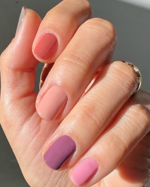 Zbliżenie na dłoń z wiosennym manicurem gradientowym (skittles) w odcieniach różu. Krótkie paznokcie, opiłowane na kształt kwadratowy, są pomalowane różnymi kolorami: od ciemnej, zgaszonej fuksji/śliwki na palcu wskazującym, przez intensywny róż i pastelowy, morelowy róż na palcach środkowym i serdecznym, aż do ciepłej, zgaszonej czerwieni/koralu na małym palcu.