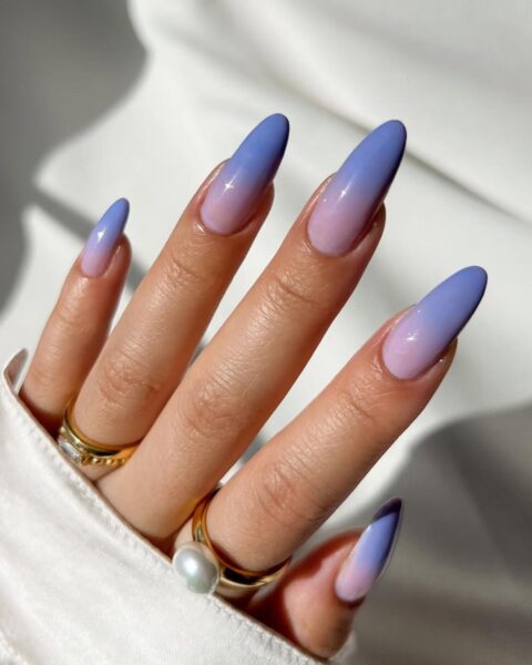 Zbliżenie na dłoń z długimi, szpiczastymi paznokciami, na których wykonano błyszczący manicure ombre (gradient). Kolory płynnie przechodzą od delikatnego, pastelowego różu/lila u nasady paznokcia, aż do nasyconego chabrowego/błękitnego fioletu na końcówkach. Dłoń nosi dwa złote pierścionki – jeden z perełką i jeden z geometrycznym kryształkiem – i jest częściowo ukryta w białym, satynowym rękawie.