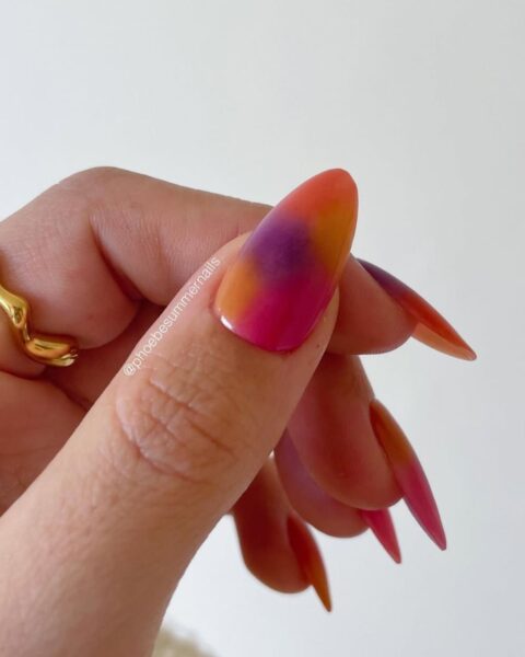 Zbliżenie na dłoń z bardzo długimi, szpiczastymi paznokciami, na których wykonano jaskrawy manicure ombre/gradient w stylu zachodu słońca. Kolory płynnie przechodzą od intensywnej fuksji/różu na końcówkach, przez pomarańcz i żółć, aż do głębokiego fioletu pośrodku płytki. Na palcu serdecznym widoczny jest złoty pierścionek o nieregularnym, falistym kształcie.