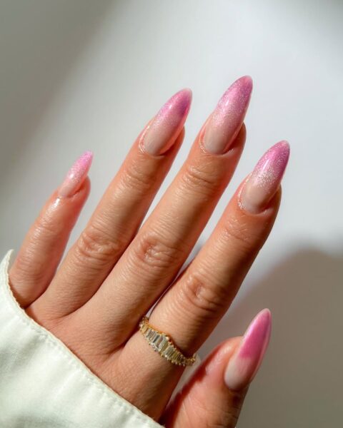 Zbliżenie na dłoń z gradient manicure na bardzo długich, migdałowych paznokciach. Paznokcie mają przezroczystą, beżową bazę, a końcówki są w kolorze intensywnego, brokatowego różu. Widoczny jest efekt cat eye (kocie oko) oraz połyskujący efekt chrome. Na palcu serdecznym widoczny jest złoty pierścionek z rzędem prostokątnych kryształków, a dłoń jest częściowo zakryta białym rękawem.