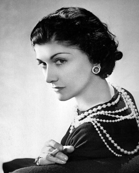 Kultowy, czarno-biały portret Coco Chanel, ikony mody. Na zdjęciu projektantka nosi swoje słynne długie sznury pereł i duże kolczyki, które symbolizują rewolucję w biżuterii. Jej spojrzenie wyraża siłę i klasę.