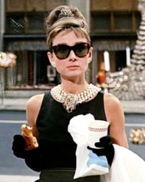 Kadr z filmu "Śniadanie u Tiffany’ego" przedstawiający Audrey Hepburn w roli Holly Golightly. Aktorka ma na sobie czarną sukienkę, duży naszyjnik, okulary przeciwsłoneczne i długie rękawiczki, trzymając w dłoniach kawę i rogalika.
