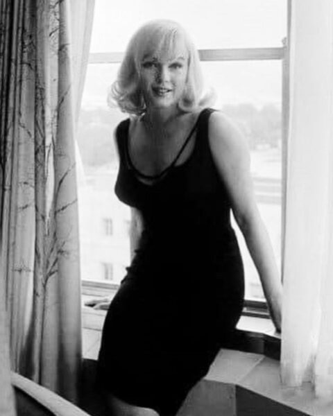 Klimatyczny, czarno-biały portret Marilyn Monroe. Gwiazda uśmiecha się lekko do kamery, siedząc w prostej, eleganckiej czarnej sukience na parapecie, wpadające przez okno światło podkreśla jej ikoniczny wizerunek.