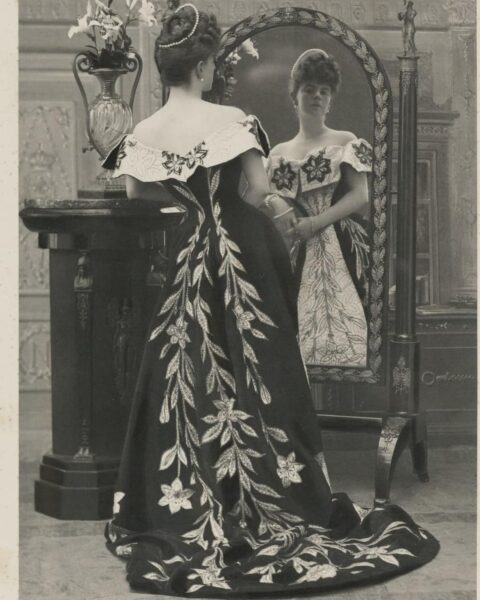 Zdjęcie kobiety w długiej sukni z kwiatowymi motywami, która przegląda się w lustrze. To zdjęcie z 1896 roku. To kreacja, której autorem jest Charles Frederick Worth, który zorganizował też pierwszy pokaz mody na świecie.