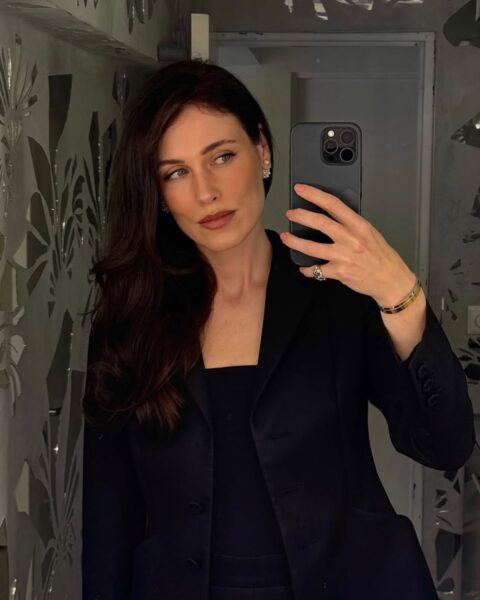 Kobieta o długich, ciemnych, lśniących włosach, ubrana w czarną marynarkę, robi sobie selfie w lustrze. Ma na sobie delikatne kolczyki i elegancką biżuterię. W tle widać lustro ozdobione wzorem z kawałków szkła.