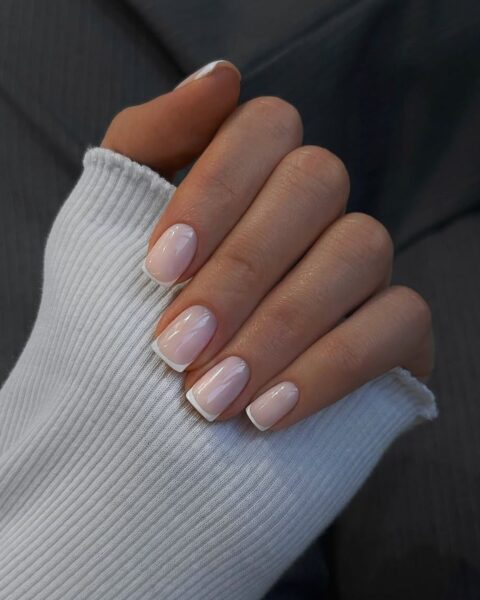 Zdjęcie dłoni ze zbliżeniem na paznokcie ze stylizacją french manicure. Paznokcie ą kwadratowe, średniej długości. Dłoń pozuje na szarym tle.