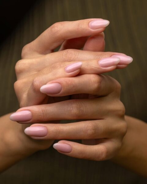 Zdjęcie dłoni ze zbliżeniem średniej długości, migdałowe paznokcie w kolorze pink nude. Dłonie są splecione i prezentują manicure. To odpowiedź na to, jakie paznokcie do pracy w biurze są odpowiednie.