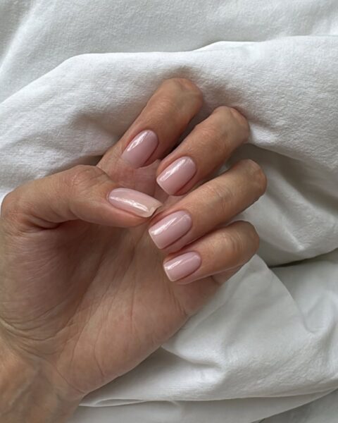 Zdjęcie dłoni ze zbliżeniem manicure do pracy: kwadratowe paznokcie w kolorze nude. Dłoń pozuje na tle białego materiału.