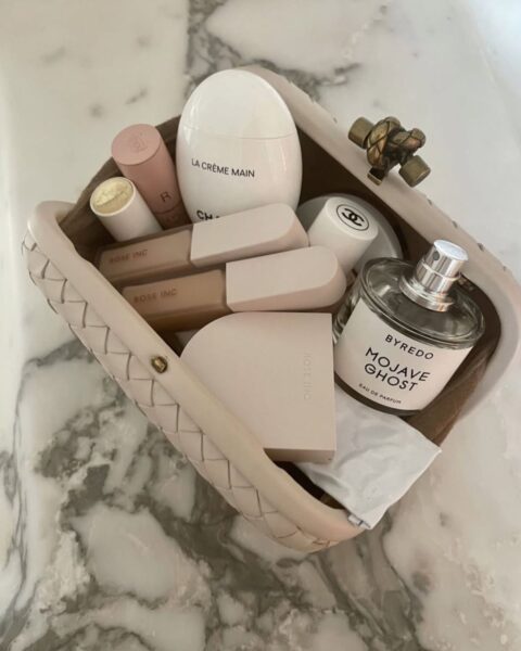Zdjęcie, na którym stoi kosmetyczka do pracy w kolorze beżowym, pleciona. Jest otwarta i widać jej wnętrze, zawartość, czyli: perfumy, pomadki, błyszczyki, pudry, krem do rąk.