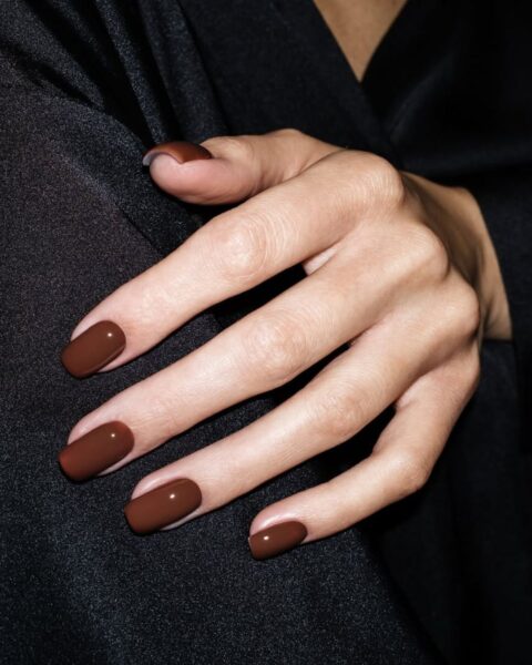 Zdjęcie ze zbliżeniem na sensualny manicure: kwadratowe, długie paznokcie w kolorze ciepłego brązu. Dłoń opiera się o ramię.