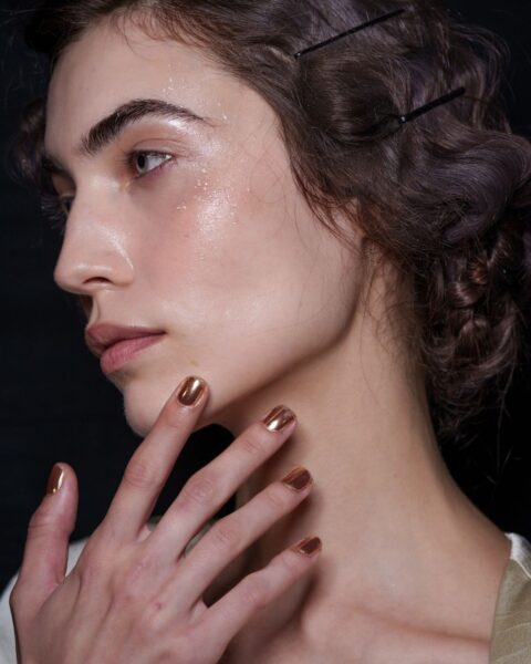 Profil modelki z fryzurą w stylu retro i efektem mokrej skóry na twarzy, prezentująca manicure w najmodniejszych kolorach z wybiegów – paznokcie pomalowane lśniącym, metalicznym lakierem w odcieniu brązu lub miedzi.