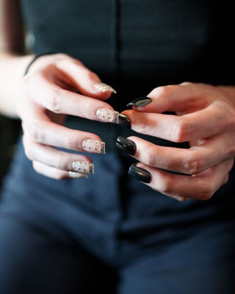 Zbliżenie na dłonie modelki z wybiegu trzymane na wysokości klatki piersiowej na tle ciemnego stroju (czarnej bluzki i granatowych spodni). Modelka prezentuje kontrastujący manicure: na lewej dłoni paznokcie są pomalowane na jasny, transparentny kolor i ozdobione drobnymi, złotymi kwadratowymi ćwiekami oraz metalicznymi końcówkami typu french, natomiast na prawej dłoni paznokcie są pomalowane na lśniący, głęboki czarny kolor.