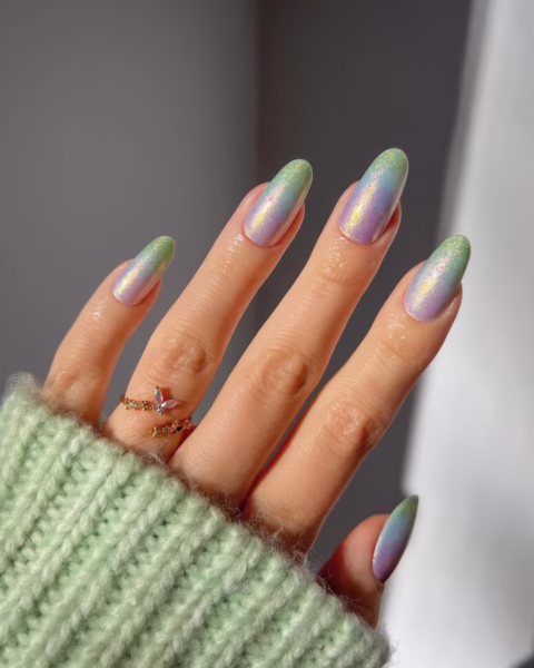 Zadbana dłoń z manicure w pełnych radości odcieniach ombre. Paznokcie, o kształcie migdałków, zmieniają kolor od jasnej zieleni u nasady, przez delikatny fiolet, aż po błękitne końcówki. Wykończenie jest błyszczące, z opalizującym efektem. Na palcu serdecznym widać delikatny, złoty pierścionek w kształcie motyla, a dłoń jest częściowo zasłonięta rękawem swetra w jasnym, pistacjowym kolorze. Tło jest stonowane, w odcieniach szarości, z wyraźnymi promieniami światła, które dodatkowo podkreślają połysk manicure.