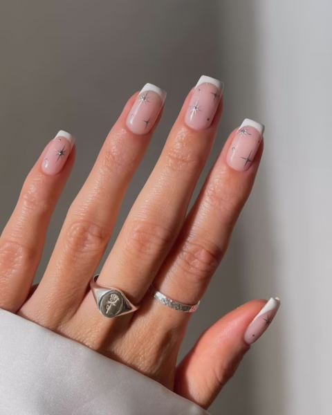 Manicure w nowoczesnym wydaniu. Paznokcie mają naturalny, lekko transparentny, beżowy odcień z białym, klasycznym french manicure. Każdy paznokieć jest ozdobiony drobnymi, lśniącymi gwiazdkami. Na palcu serdecznym widoczne są dwa srebrne pierścionki, z czego jeden to sygnet z grawerowaną różą. Dłoń spoczywa na białym materiale, który prawdopodobnie jest rękawem koszuli. Tło jest jasne, jednobarwne.