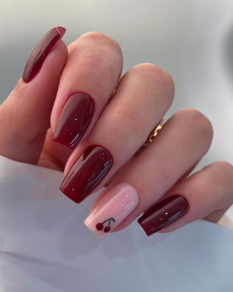 Kobiece dłonie z paznokciami pomalowanymi w odcieniu głębokiej wiśni. Dwa z nich są w jednolitym kolorze, a jeden, na palcu serdecznym, ma french manicure z subtelnym, beżowym tłem i akcentem dwóch małych, namalowanych wisienek. Paznokcie są w kształcie prostokąta, z wyraźnym, błyszczącym wykończeniem. W tle widać jasny, biały materiał, a dłoń jest częściowo zasłonięta.