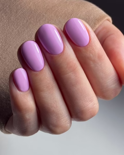 Zadbane, kobiece dłonie z manicure w pastelowym odcieniu fioletu, wpadającym w liliowy róż. Paznokcie mają owalny kształt, a na skórze widać subtelny błysk, sugerujący, że była ona nawilżana. W tle widoczny jest beżowy materiał, który prawdopodobnie jest rękawem swetra.