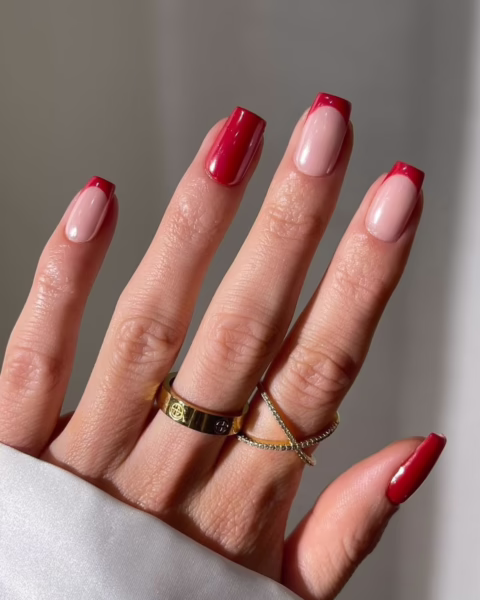 Manicure, który łączy klasyczną, francuską wersję z nowoczesnym akcentem. Dwa paznokcie są pomalowane na jednolity, intensywny odcień wiśniowej czerwieni, podczas gdy pozostałe mają naturalny, beżowy kolor z czerwoną końcówką, przypominającą french manicure. Na palcu serdecznym widać dwa złote pierścionki, z czego jeden jest cienki, a drugi grubszy. Tło jest stonowane, a dłoń rzuca delikatny cień na ścianę.
