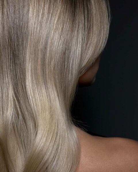 Zdjęcie kobiety stojącej tyłem. Ma blond włosy, długie, rozpuszczone na ramiona. Są bardzo gładkie, bo używa maski dociążające włosy.