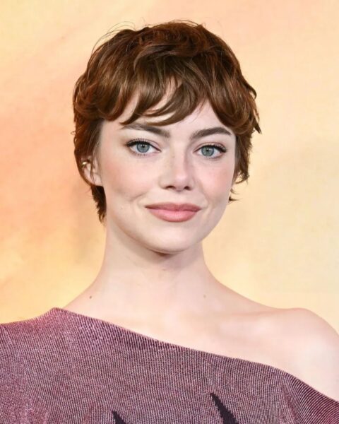 Aktorka Emma Stone w ujęciu portretowym, prezentująca krótką, rudą fryzurę typu pixie cut z naturalnie pofalowaną grzywką. Ma na sobie top lub sweter z dekoltem opadającym na jedno ramię w kolorze przygaszonego fioletu z metalicznym połyskiem. Jej makijaż jest naturalny, z delikatnym różem na policzkach i akcentem na wyraziste, niebieskie oczy. Tło jest rozmyte, w ciepłych, żółto-pomarańczowych barwach.