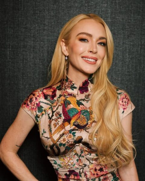 Lindsay Lohan w kolorowej, jedwabnej mini-sukience w stylu chińskim cheongsam (qipao), z wysokim kołnierzykiem (stójką) i krótkimi rękawami. Sukienka jest dopasowana w talii i pokryta bogatym wzorem w orientalne ptaki i kwiaty. Aktorka ma długie, falowane blond włosy, rozpuszczone i zaczesane na bok. Uśmiecha się, trzymając dłoń na biodrze. Ma na sobie delikatną złotą biżuterię. Tło jest ciemne i teksturowane.
