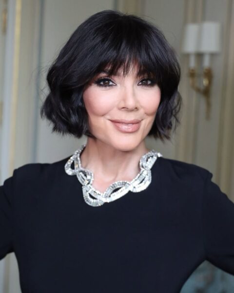 Portret uśmiechniętej Kris Jenner. Ma na sobie czarną sukienkę z długim rękawem. Uwagę zwraca masywny, srebrny naszyjnik, ozdobiony kryształkami. Włosy ma ułożone w krótkiego boba z grzywką. Makijaż oczu jest wyrazisty, w stylu smoky eye z brązowymi cieniami, a usta ma pomalowane jasną szminką. Tło jest jasne, widać na nim fragment eleganckiego żyrandola lub lampy.