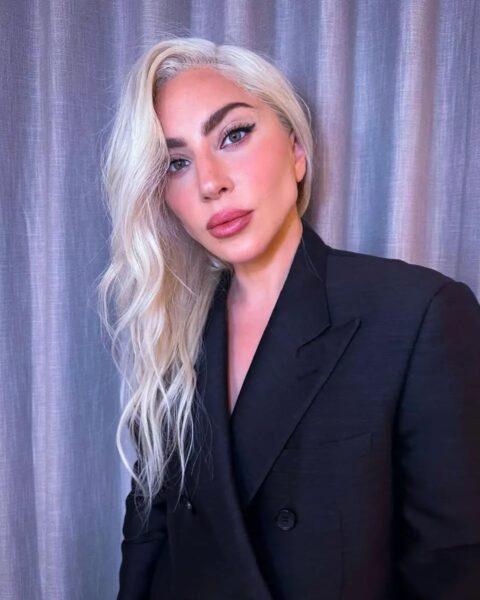 Lady Gaga w eleganckiej, czarnej, dwurzędowej marynarce o męskim kroju i podniesionych ramionach. Piosenkarka ma platynowy blond, długie, falowane włosy zaczesane na bok i wyrazisty makijaż oczu typu "smoky eye" oraz matowe, różowe usta. Zdjęcie z bliska wizerunku ikony stylu.