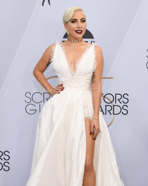 Lady Gaga na czerwonym dywanie w dramatycznej, białej sukni Dior Couture. Kreacja ma głęboki, plisowany dekolt w kształcie litery V obszyty delikatnymi frędzlami, odcinaną talię i rozcięcie eksponujące nogę. Suknię dopełniają białe szpilki, złote bransoletki i gruby, złoty choker. Włosy są ułożone w gładki, platynowy bob, a na ustach ma ciemną, burgundową szminkę. Pozuje z dłonią na biodrze na tle z logotypami Screen Actors Guild Awards.