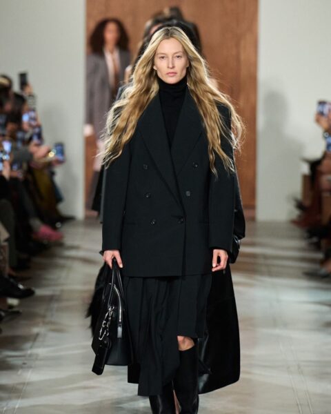 Zdjęcie z pokazu jesiennego Michaela Korsa: blond modelka z długimi włosami, ubrana w czarną marynarkę oversize, spódnicę midi w kolorze czarnym, kozaki, torebkę, idzie po wybiegu.