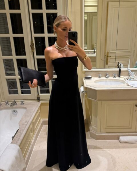 Kobieta w eleganckiej, czarnej sukni maxi bez ramiączek (strapless) robi sobie zdjęcie w lustrze łazienki hotelowej. Suknia jest uzupełniona kilkoma sznurami klasycznych pereł i złotymi bransoletkami. W ręku trzyma czarną kopertówkę, a za nią widać mężczyznę w białej koszuli. Stylizacja to kwintesencja ponadczasowej elegancji. Fraza: moda wieczorowa jak wybrać kreację na czerwony dywan.