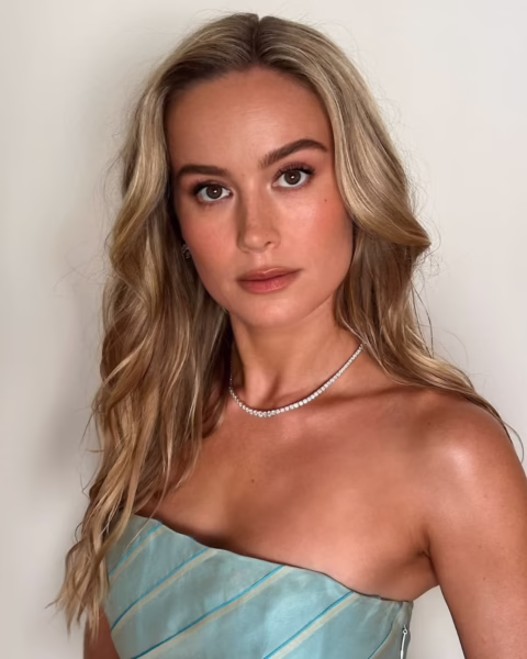 Portret Brie Larson z długimi, falowanymi włosami w odcieniu blond, w błękitnej sukience bez ramiączek i z naszyjnikiem na szyi. Patrzy prosto w obiektyw.