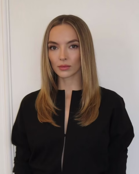 Portret Jodie Comer z prostymi, długimi włosami w odcieniu blond, w czarnej bluzce i ze złotym paskiem. Spogląda wprost na obiektyw.