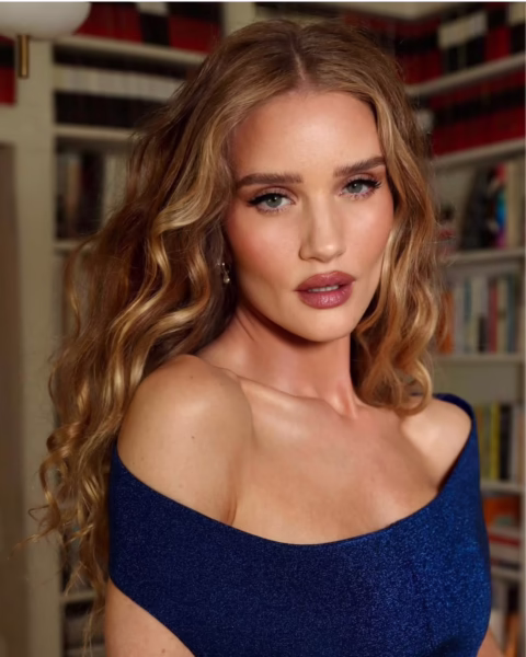 Modelka Rosie Huntington-Whiteley z długimi, blond włosami, ułożonymi w delikatne fale, ubrana w granatową sukienkę z dekoltem odsłaniającym ramiona. Tło stanowią regały z książkami.