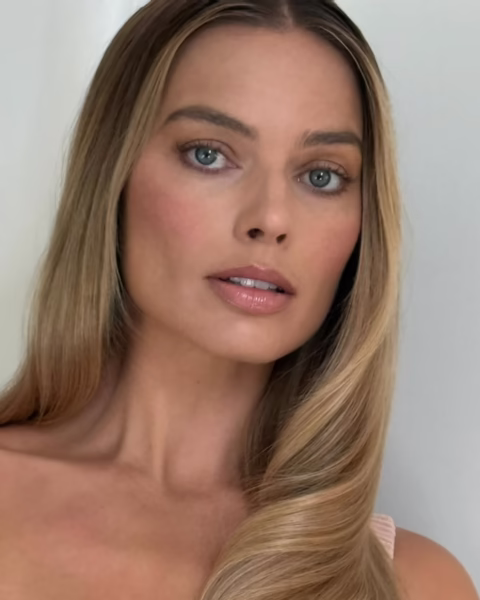 Portret Margot Robbie z długimi, blond włosami ułożonymi w delikatne fale, która patrzy prosto w obiektyw.