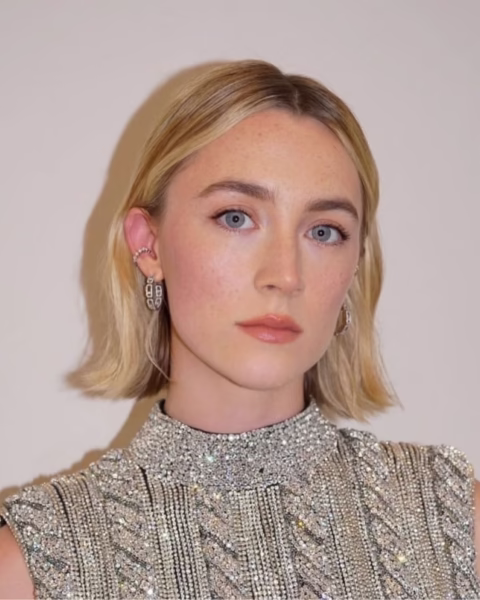 Portret aktorki Saoirse Ronan, która ma krótkie, blond włosy w stylu bob z widocznym odrostem, wpisującym się w trend recession blonde. Ma na sobie błyszczącą, wyszywaną kamieniami sukienkę, która jest idealnym uzupełnieniem jej minimalistycznego looku.