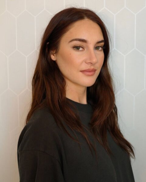Zdjęcie kobiety z długimi, brązowymi włosami w odcieni borówkowym, czyli Berry-stained brunette. Jest ubrana w czarną bluzę. Pozuje na tle szarej ściany.