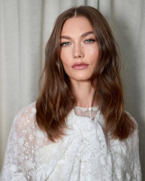 Zdjęcie kobiety z długimi, rozpuszczonymi włosami w kolorze warm beige brunette, czyli beżowego brązu. Jest ubrana w koronkową, białą bluzkę. Pozuje na tle kremowej zasłony.