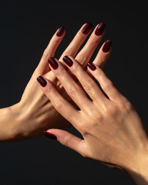 Zdjęcie dłoni z długimi, kwadratowymi paznokciami w kolorze burgundowym. Dłonie splatają się i pozują tak, by prezentować manicure. Pozują na czarnym tle.