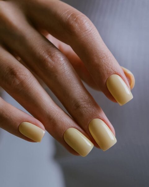 Zdjęcie dłoni prezentujące modne kolory manicure na jesień 2025: tutaj jest maślany żółty. Paznokcie są kwadratowe, średniej długości.