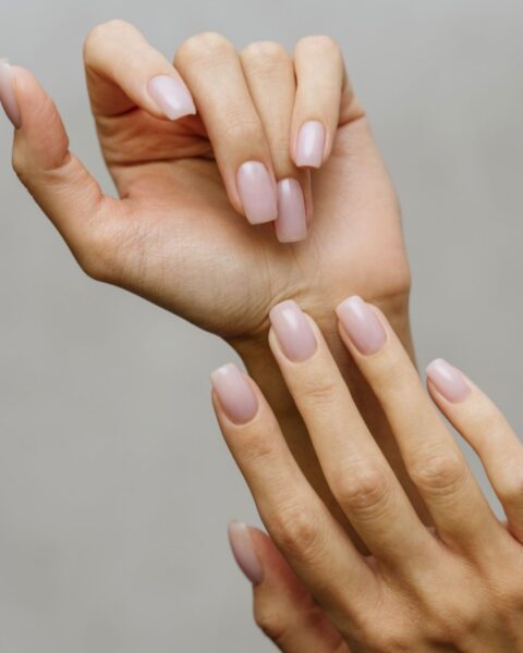 Zdjęcie dłoni z długimi, kwadratowymi paznokciami. Są pomalowane na kolor jasnego nude. Dłonie pozują tak, by prezentować manicure. Pozują na szarym tle.